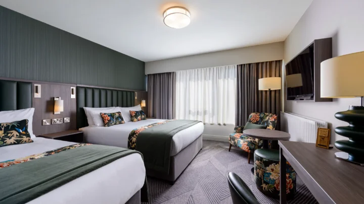 Stylish hotel room with two double beds, plush pillows, warm lighting, floral chairs, modern TV, creating a cosy retreat.