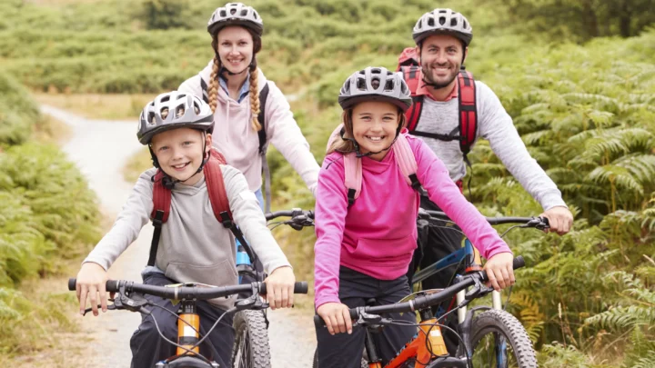 A family of four wearing helmets and backpacks rides mountain bikes on a gravel path through green countryside.