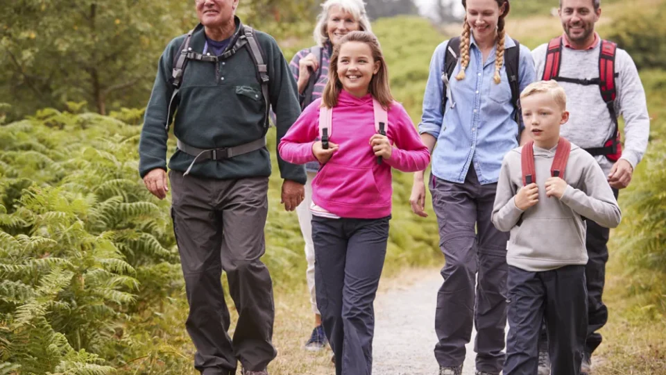 A group of six people, including adults and children, walk together on a forest trail, wearing hiking clothes and backpacks.