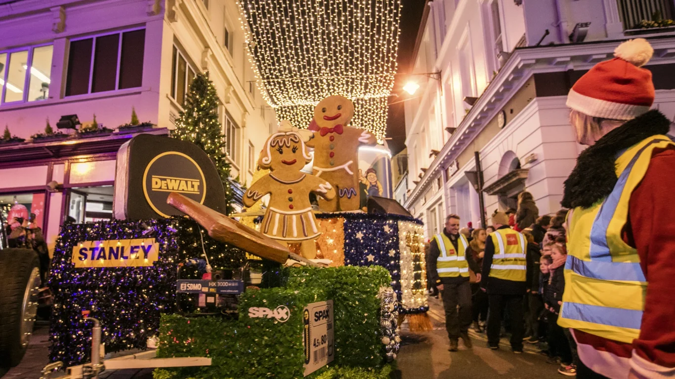 Festive street parade with gingerbread decorations and lively crowd under twinkling lights, creating a warm holiday atmosphere.