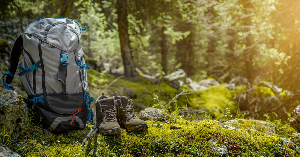 A hiking backpack, boots, and trekking poles rest on mossy ground in a sunlit forest clearing—gear up for adventure with our Spring Offer.