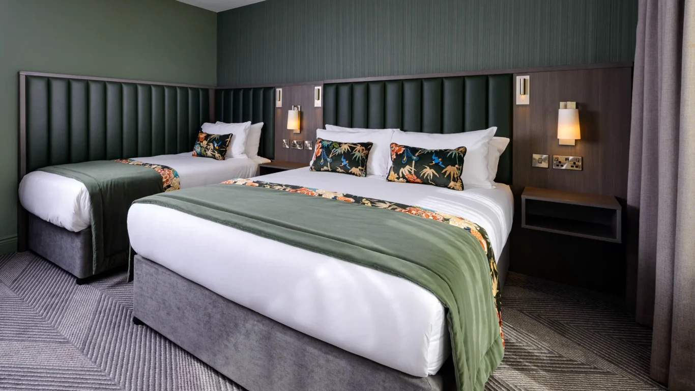 Modern hotel room with two plush double beds, green accents, stylish patterned cushions, and ambient lighting.