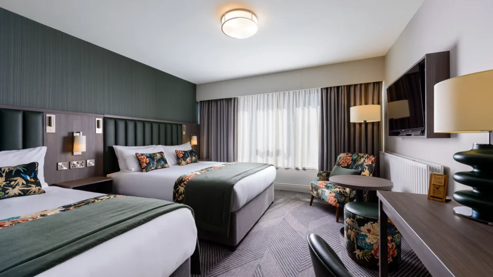 Spacious hotel room with two double beds, stylish decor, armchair, large TV, and natural light for a relaxing stay.