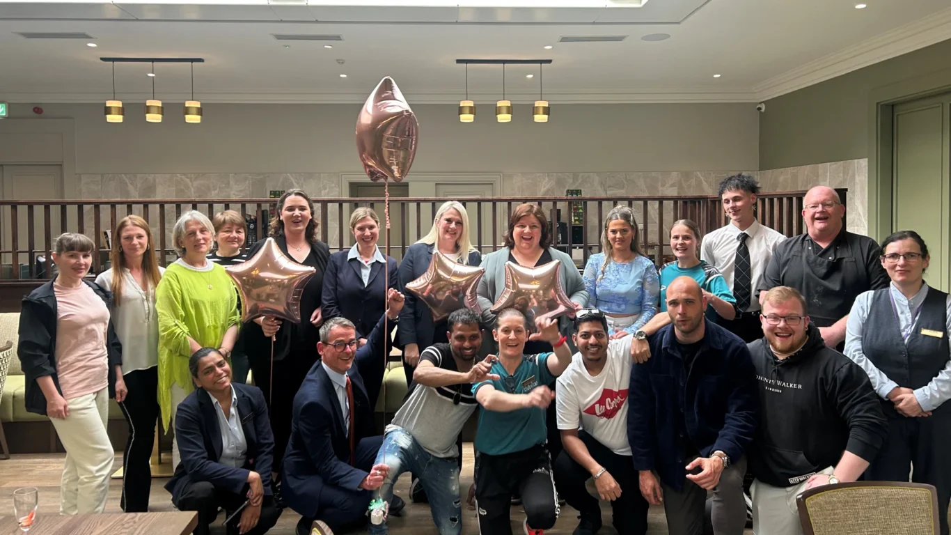 A group of people pose indoors for a photo, some holding metallic star-shaped balloons. They are dressed in a mix of smart and casual clothing.