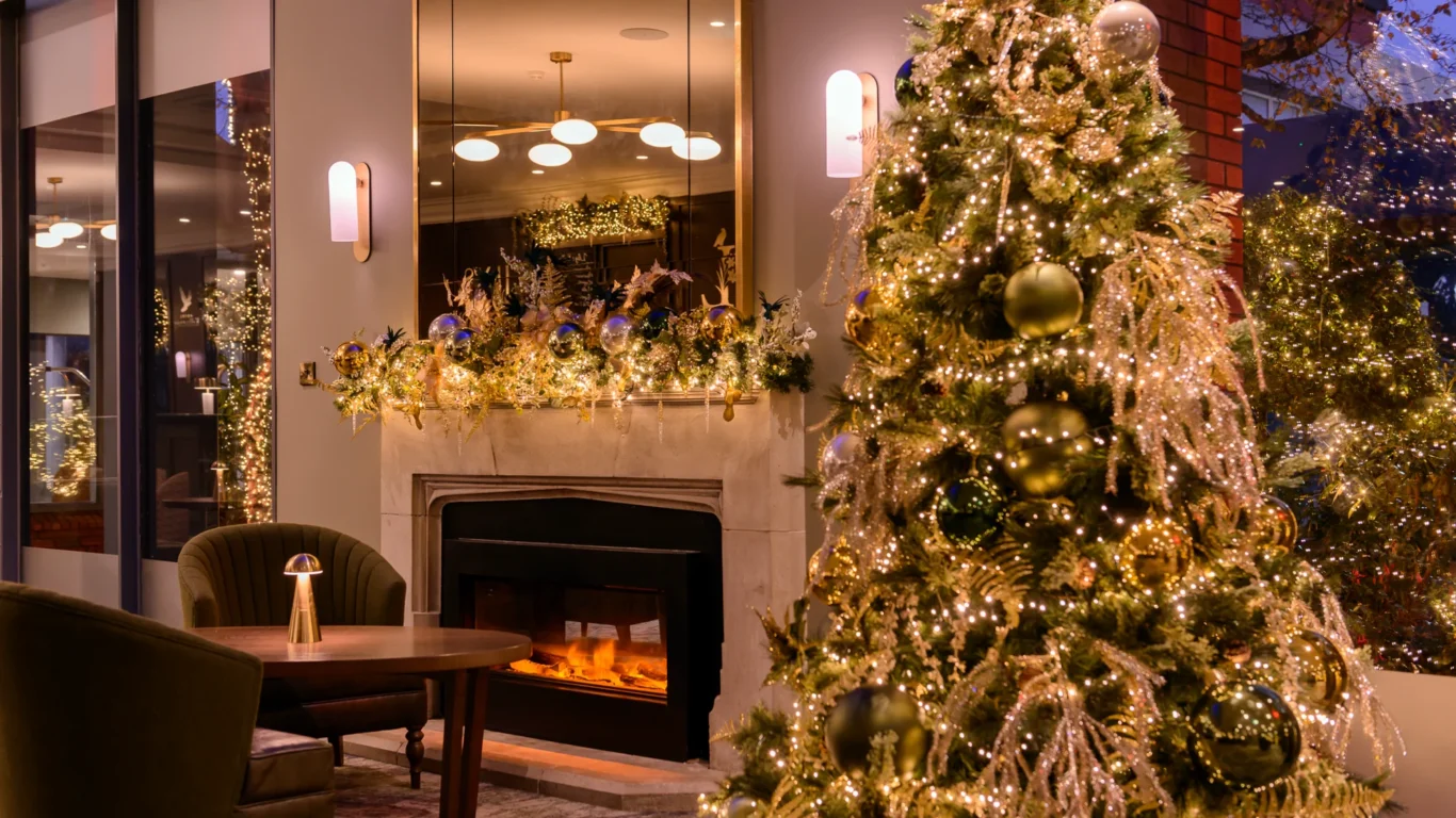 A decorated Christmas tree with lights stands near a lit fireplace in a modern living room adorned with festive garland and ornaments.