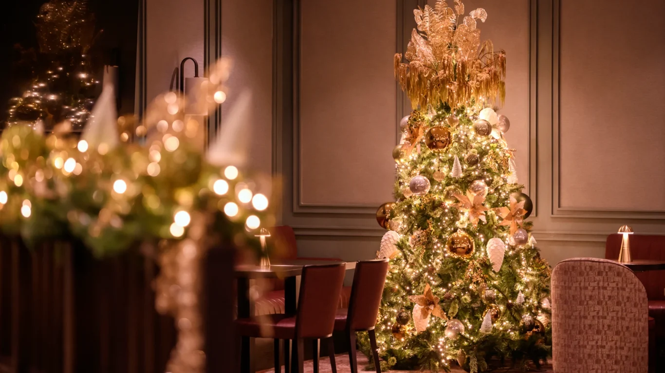 A decorated Christmas tree with gold ornaments and lights stands in an elegant, softly lit room with chairs and tables.