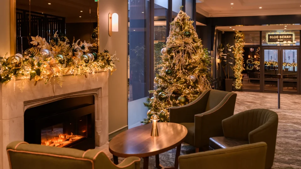 A hotel lounge decorated for Christmas with a lit fireplace, garland, a large decorated Christmas tree, and comfortable seating near large windows.