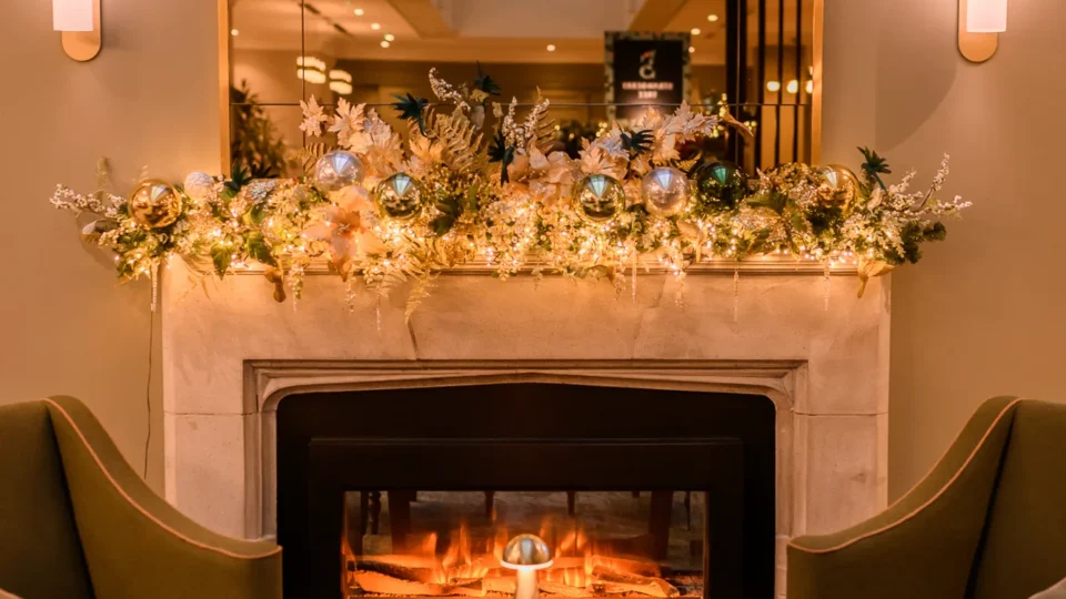 Four green armchairs and a round wooden table are arranged around a fireplace decorated with flowers and string lights under a large mirror.