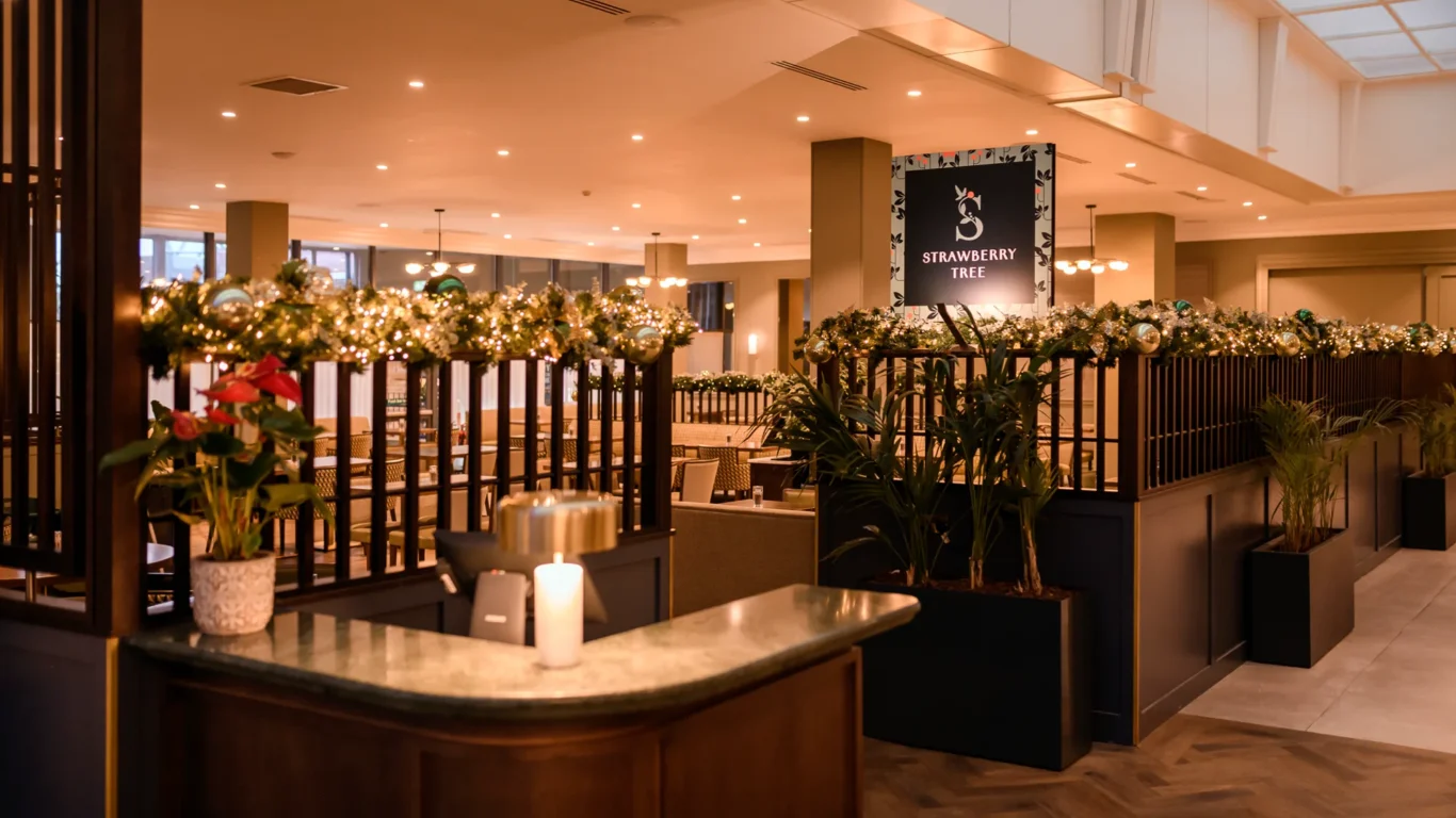 Reception area of a restaurant with festive garlands on railings, potted plants, a lit candle, and a sign reading 