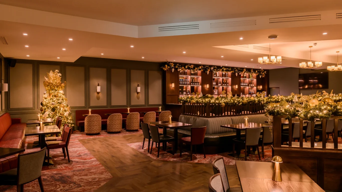 A warmly lit restaurant bar decorated with festive garlands and Christmas lights, featuring wooden tables, cushioned chairs, and a well-stocked bar area.