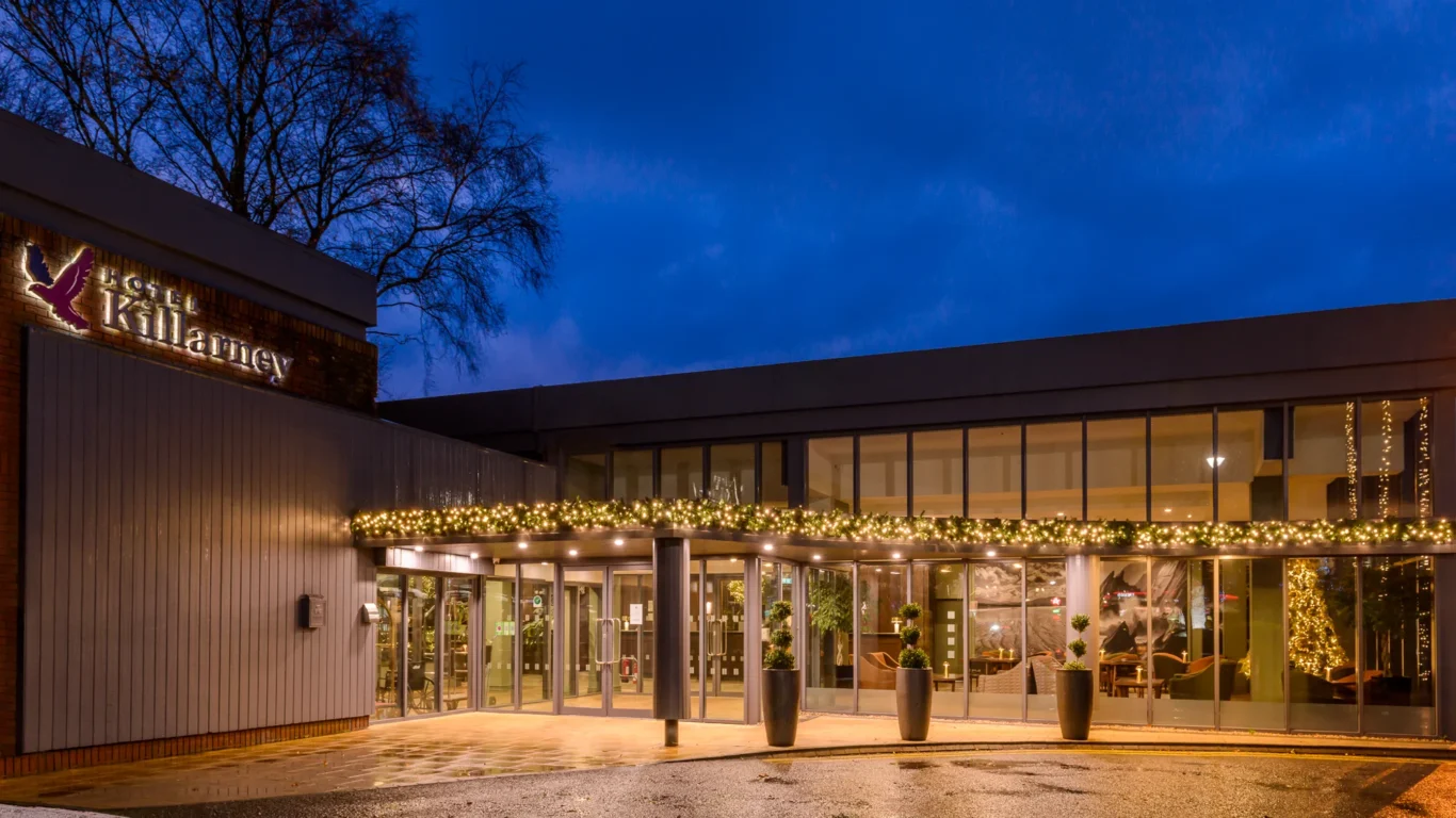 The exterior of a modern hotel building at dusk, decorated with string lights and holiday garlands, with glass doors and large windows.