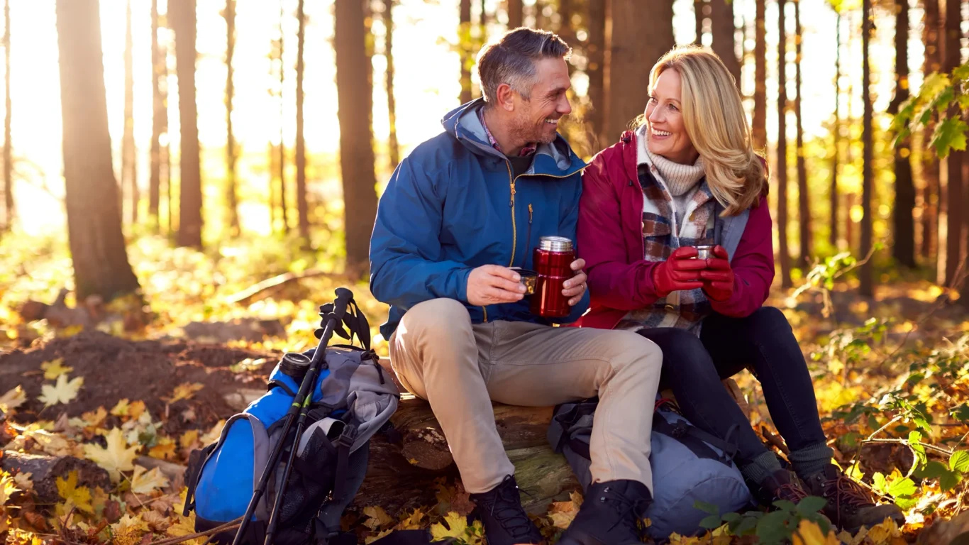 Couple enjoying a warm drink in a sunlit autumn forest, surrounded by hiking gear, embodying outdoor adventure and relaxation.